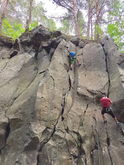 Twee klimmers beklimmen een steile rotswand in een bos. Omringd door bomen tonen ze hun klimvaardigheden in de natuur.