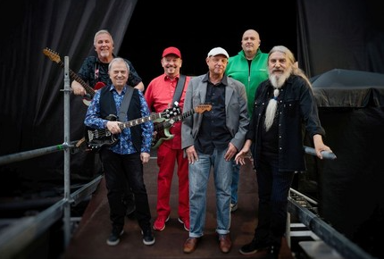 Sechs Musiker stehen zusammen auf einer Bühne. Sie tragen verschiedene bunte Outfits und halten Instrumente in der Hand.