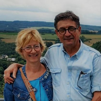 A smiling couple poses in front of a picturesque landscape. In the background, green hills and a cloudy sky can be seen.
