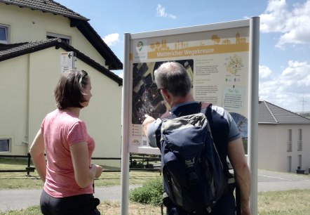 Twee wandelaars staan voor een wandelbord met de tekst 'Metterich wayside crosses' en krijgen informatie over de route. Op de achtergrond zijn huizen te zien., © Tourist-Information Bitburger Land