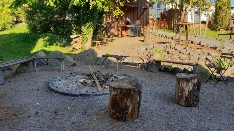 Un endroit confortable autour d'un feu de camp avec des bûches et des bancs. Entouré d'espaces verts et d'un bâtiment rustique à l'arrière-plan.