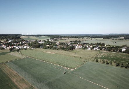 Vue aérienne d'Idenheim : une agglomération rurale entourée de champs verdoyants, avec des éoliennes à l'horizon sous un ciel dégagé., © Ingrid Penning