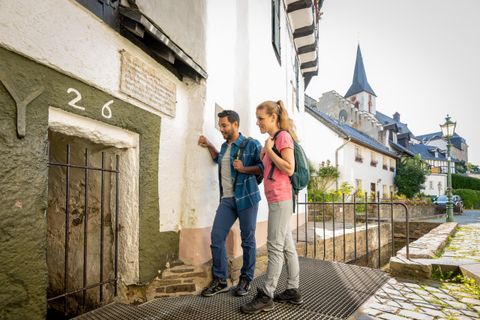 Een paar staat voor een oud, historisch gebouw met een kleine deur. Op de achtergrond zijn pittoreske vakwerkhuizen en een kerktoren te zien.