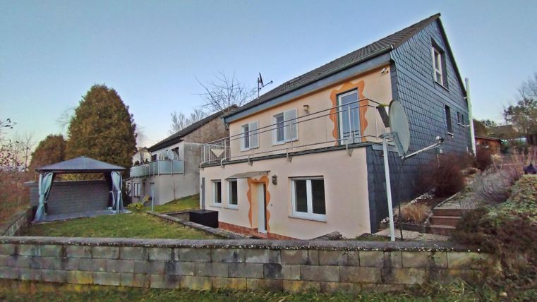 Une maison moderne avec un jardin et une petite terrasse. En arrière-plan, des arbres sont visibles.