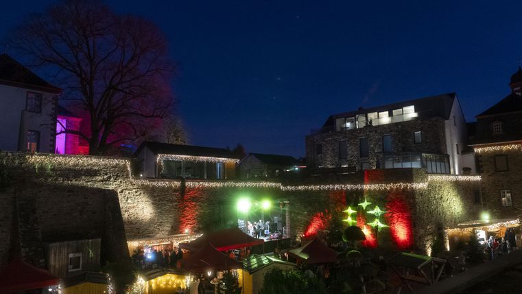 Eine Gruppe von Menschen prostet sich mit Getränken zu. Im Hintergrund ist ein festlich dekorierter Stand mit dem Schild "HAUS BACHTAL" zu sehen.