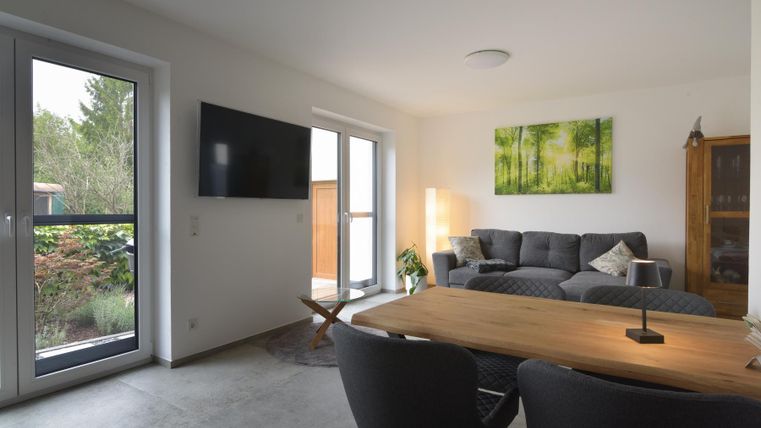 Ein modernes Wohnzimmer mit einem grauen Sofa und einem Holztisch. Große Fenster lassen viel Licht herein und ein Kunstwerk mit grünen Bäumen schmückt die Wand.