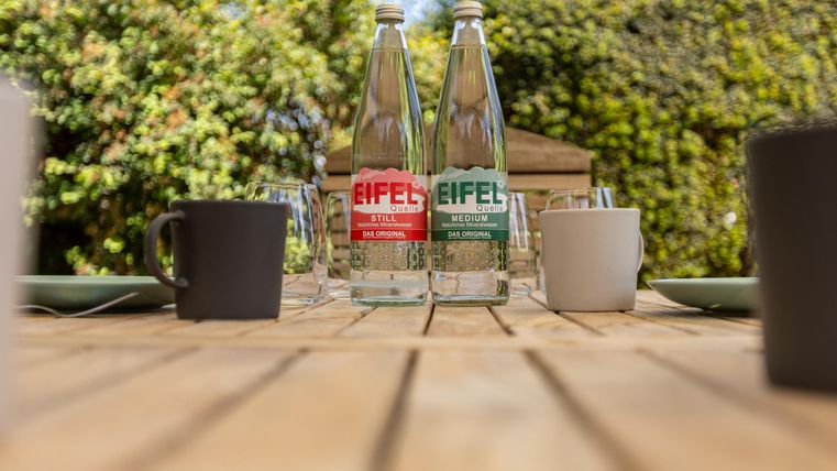 Twee flessen water met labels in rood en groen staan op een houten tafel. Om hen heen zijn glazen en kopjes geplaatst op een mooie, groene achtergrond.