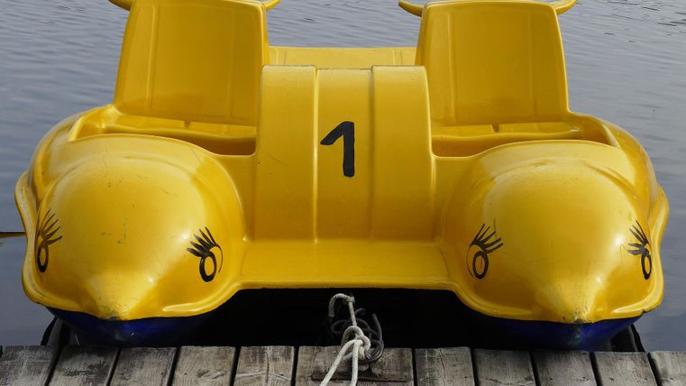 A yellow pedal boat with two seats is floating on the water and is tied to a wooden jetty with a cord.