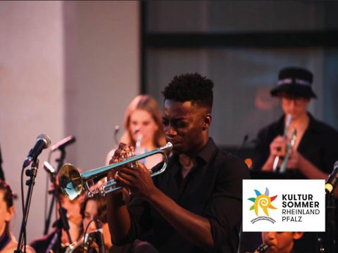 Zwei Musiker stehen auf einer Bühne und lächeln in die Kamera. Beide halten Akustikgitarren, während im Hintergrund eine Band zu sehen ist.