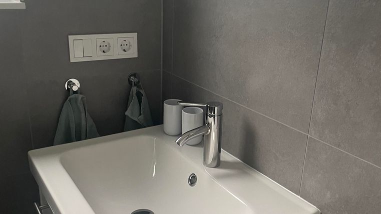 Ein modernes Badezimmer mit einem kleinen Waschbecken und eleganten Armaturen. Die Wände sind mit grauen Fliesen verkleidet und ein Fenster sorgt für Tageslicht.