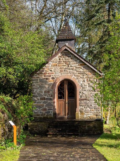 A small stone chapel surrounded by green foliage and trees. The entrance has an elegant wooden door and the architecture is simple and charming.