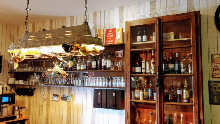 A cozy bar area with a wooden shelf wall. The counter is decorated with glasses and a selection of spirits.