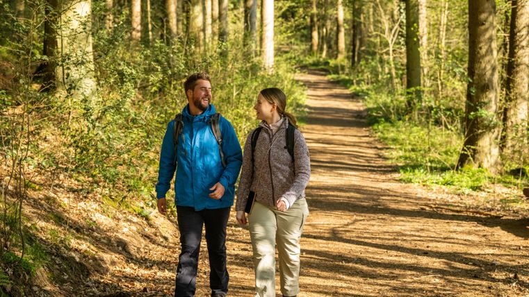 Een paar wandelt langs een schilderachtig bospad. De zon schijnt door de bomen en creëert een uitnodigende sfeer.