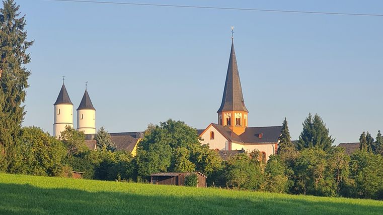 Eine malerische Landschaft mit einer Kirche und zwei Türmen im Hintergrund. Umgeben von grünen Wiesen und einem klaren blauen Himmel.