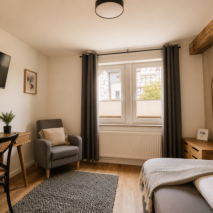 A bright room with a cozy armchair, a table, and a television. Large windows provide plenty of daylight and a view of the outdoors.