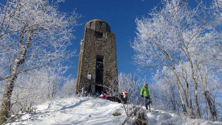 Ein schneebedeckter Berg mit einem alten Turm und frostigen Bäumen. Ein Wanderer in grüner Kleidung steht in der Nähe des Turms.