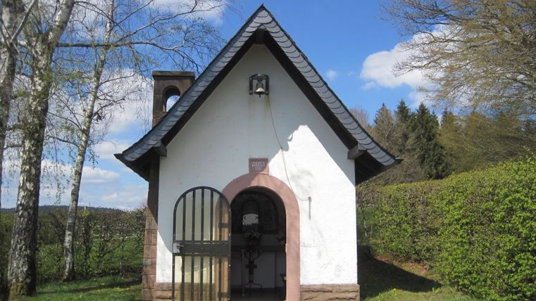 Petite chapelle blanche avec un toit noir dans un environnement rural, entourée d'arbres et de haies.