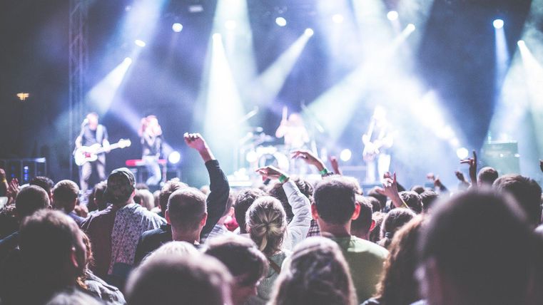 Ein Publikum genießt ein Live-Konzert mit einer Band auf der Bühne. Bunte Lichter und eine energiegeladene Stimmung prägen die Szene.