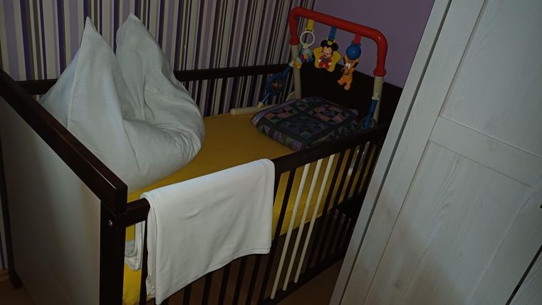 Un lit d'enfant avec un matelas jaune et des coussins. Sur le côté, des jouets colorés pendent.