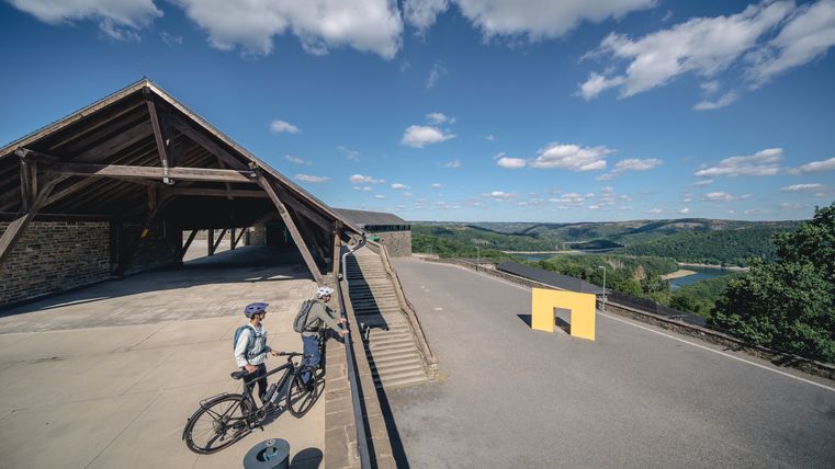 Two cyclists stand in front of a building with a wooden roof and look out over a hilly landscape with a river.