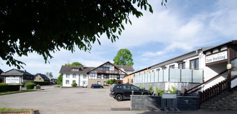 Een rustige parkeerplaats voor een hotel met meerdere gebouwen. De lucht is helder en zonnig, omringd door bomen.