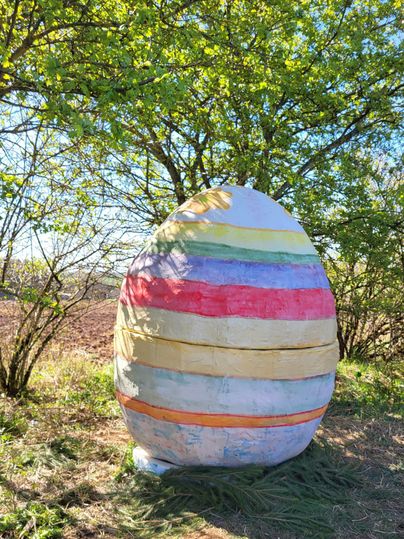 Een grote, kleurrijk beschilderde paasei staat buiten, omringd door bomen. De lucht is helder en het is mooi zonnig.