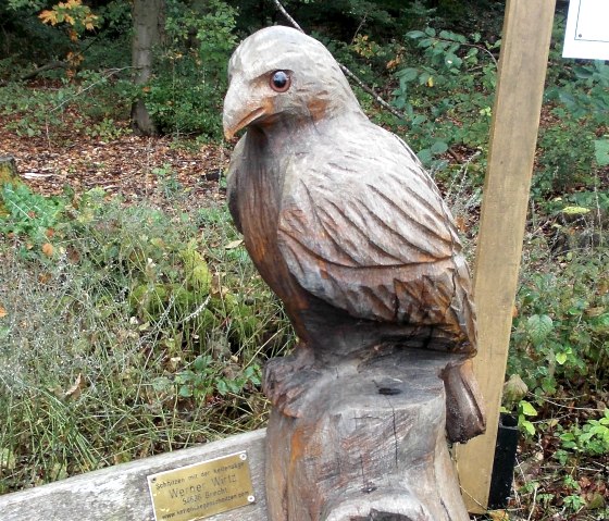 Eine Holzskulptur eines Vogels sitzt auf einem Baumstamm im Wald. Eine Plakette ist an der Skulptur angebracht., © Tourist-Information Bitburger Land