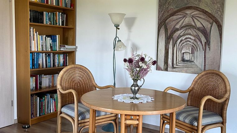 Ein gemütlicher Raum mit einem runden Tisch und zwei Stühlen. Im Hintergrund steht ein Bücherregal und ein Gemälde ist an der Wand.