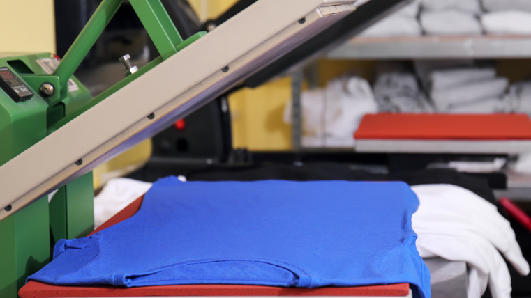A blue T-shirt is lying in a fabric press. In the background, you can see a shelf with folded white T-shirts.