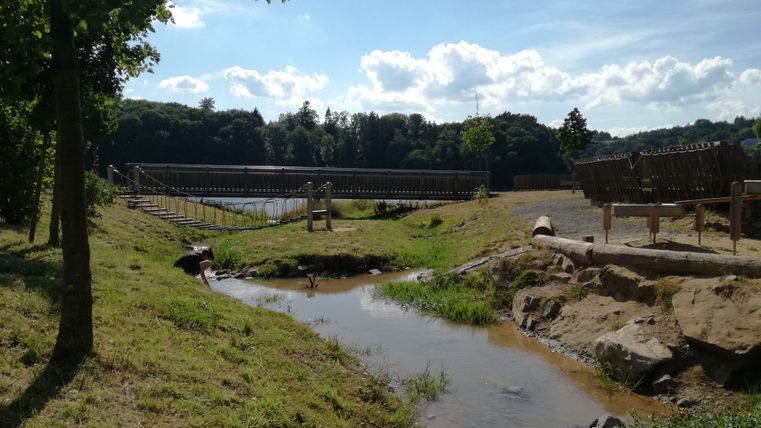 Ein kleiner Bach fließt durch eine grüne Landschaft mit einer Holzbrücke im Hintergrund.