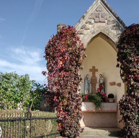A small chapel in Wolsfeld, surrounded by red climbing plants. Religious statues can be seen inside. The sky is clear and blue., © Tourist-Information Bitburger Land