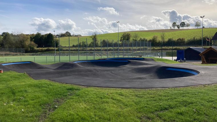 Ein Spielplatz mit einer wellenförmigen Gummibahn und Rasenflächen. Im Hintergrund sind Tennisplätze und ein grüner Hügel zu sehen.