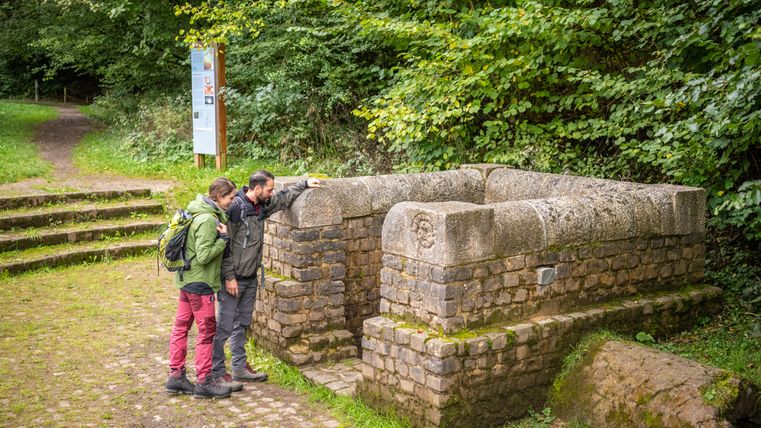 Deux personnes regardent une fontaine historique dans un environnement forestier.