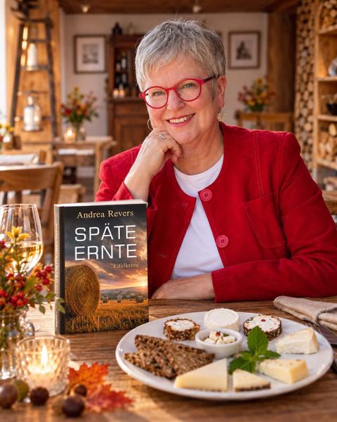 Eine lächelnde Frau sitzt an einem Tisch mit einer Auswahl an Käse und Brot. Auf dem Tisch liegt das Buch "Späte Ernte" von Andrea Revers.