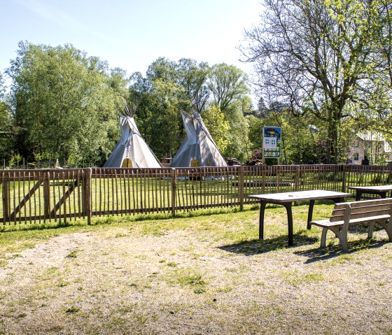 Une aire de jeux avec deux tipis derrière une clôture en bois, entourée d'arbres. Au premier plan, des tables et des bancs sur un sol en gravier., © Tourist-Information Bitburger Land_M. Mayer