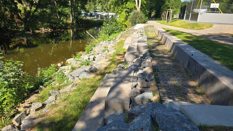 A narrow path along a body of water with stones and green areas. In the background, trees and a sports field are visible.
