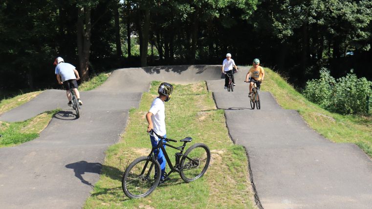 Children are riding on a BMX park with hilly tracks. The sun is shining and the surroundings are green.