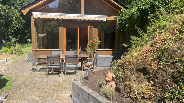 A cozy wooden house with large windows and a terrace. The garden is green and well-maintained, surrounded by plants and a stone path.