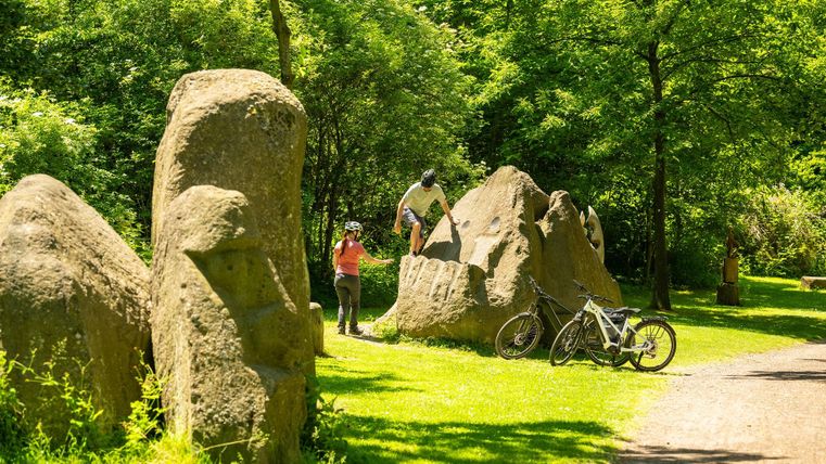 A green park landscape with large rocks and cyclists. In the background, there are two people exploring the rocks.