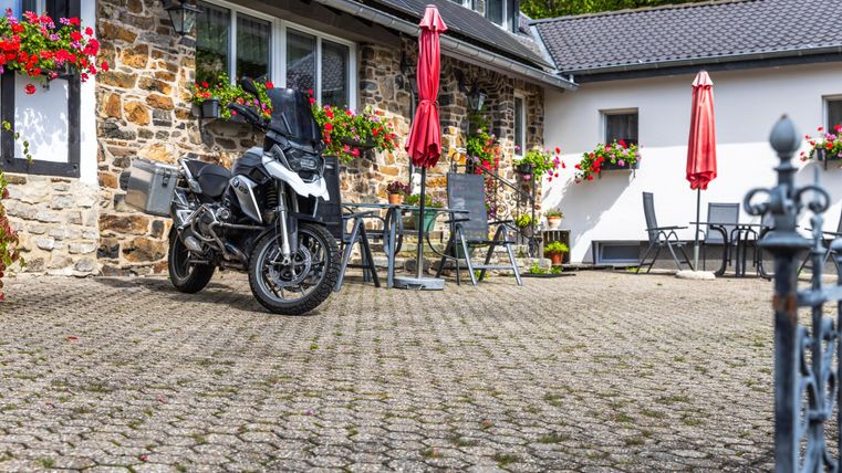 A motorcycle stands in a paved courtyard in front of a beautiful house. Red umbrellas and flower boxes give the surroundings an inviting atmosphere.