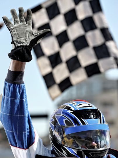 Ein Rennfahrer hebt die Hand mit einer schwarz-weißen Schachflagge. Im Hintergrund ist eine Rennstrecke zu sehen.