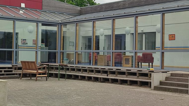 Un complexe immobilier moderne avec de grandes fenêtres en verre et une zone couverte. Devant le bâtiment, des chaises en bois sont visibles sur une surface pavée.