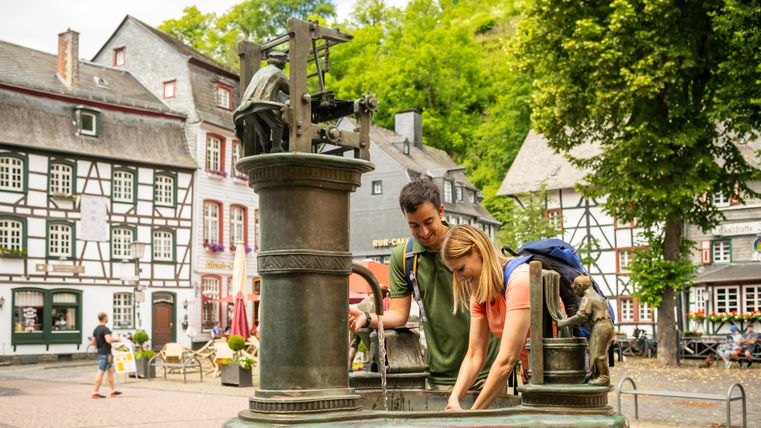Twee mensen verfrissen zich bij de Tuchmacherbrunnen fontein in Monschau, omringd door vakwerkhuizen en bomen.