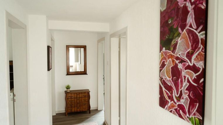 Un couloir lumineux avec des murs blancs et un grand miroir.
Sur le mur, il y a une peinture colorée et il y a une petite commode en bois.