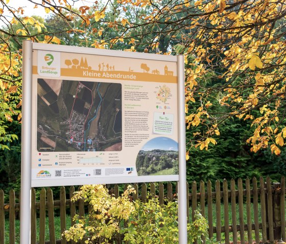 Sign for the 'Kleine Abendrunde' hiking route in Bitburger Land, surrounded by autumnal trees and a wooden fence.