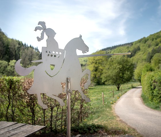 Metal figure of a knight on a horse with a lance, in front of a green landscape and a path. Trees and a blue sky can be seen in the background., © Tourist-Information Bitburger Land_Monika Mayer