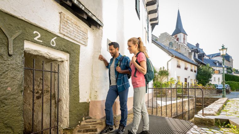 Een paar staat voor een oud, historisch gebouw met een kleine deur. Op de achtergrond zijn pittoreske vakwerkhuizen en een kerktoren te zien.