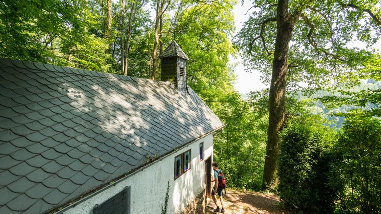 Een wandelaar betreedt een kleine hermitage in het bos met een leistenen dak en klokkentoren.