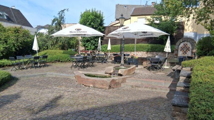 Un espace extérieur confortable avec des tables et des chaises sous des parasols. Entouré de plantes vertes et d'une façade en pierre pittoresque.