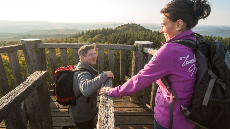 Deux personnes sur une tour d'observation avec vue sur des collines boisées en arrière-plan.